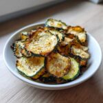 A white bowl filled with golden brown, seasoned Crunchy Zucchini Chips, ready for snacking.