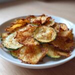 A close-up of a white bowl filled with golden brown, seasoned Crunchy Zucchini Chips.