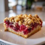 A close-up of a square slice of delicious raspberry crumb bars with a thick layer of bright red filling and a golden crumble topping.
