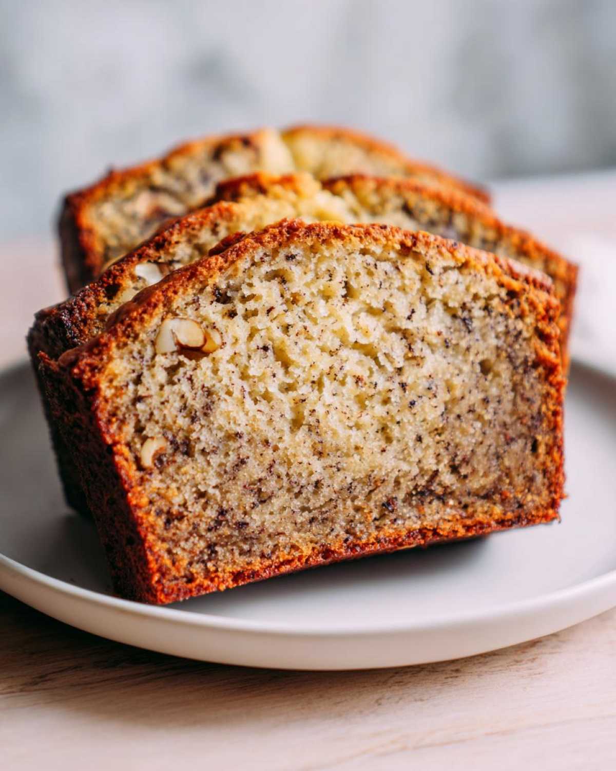 Close-up of three slices of moist Favorite Banana Bread showing nuts and dark banana specks on a white plate.