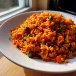 A close-up of fluffy Authentic Mexican Rice, bright orange-red, mixed with green peas and diced carrots on a white plate.