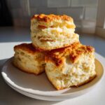 Three tall, flaky Buttermilk Biscuits stacked on a rustic plate, showing golden tops and airy interior.