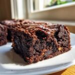 A close-up of a rich, fudgy square of Banana Bread Brownies with a shiny, cracked top, sitting on a white plate.