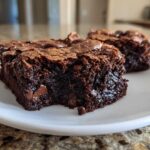 Two rich, fudgy brownie squares with a crackly top and visible chocolate chips served on a white plate.