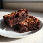 Two rich, fudgy brownie squares stacked slightly on a white plate, showing a shiny, cracked top crust.
