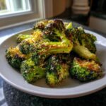 A close-up of roasted Garlic Parmesan Broccoli florets piled high on a white plate, showing crispy edges and grated cheese.