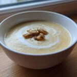 A white bowl of creamy yellow Garlic Soup with Roasted Bulb pieces and a swirl of cream.