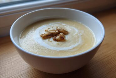 A white bowl of creamy yellow Garlic Soup with Roasted Bulb pieces and a swirl of cream.