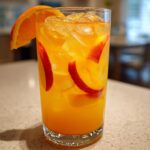 Tall glass filled with bright orange Ginger Peach Lemonade, ice, peach slices, and an orange wedge garnish.