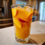 Close-up of a tall glass filled with vibrant orange Ginger Peach Lemonade, ice cubes, and fresh peach slices.