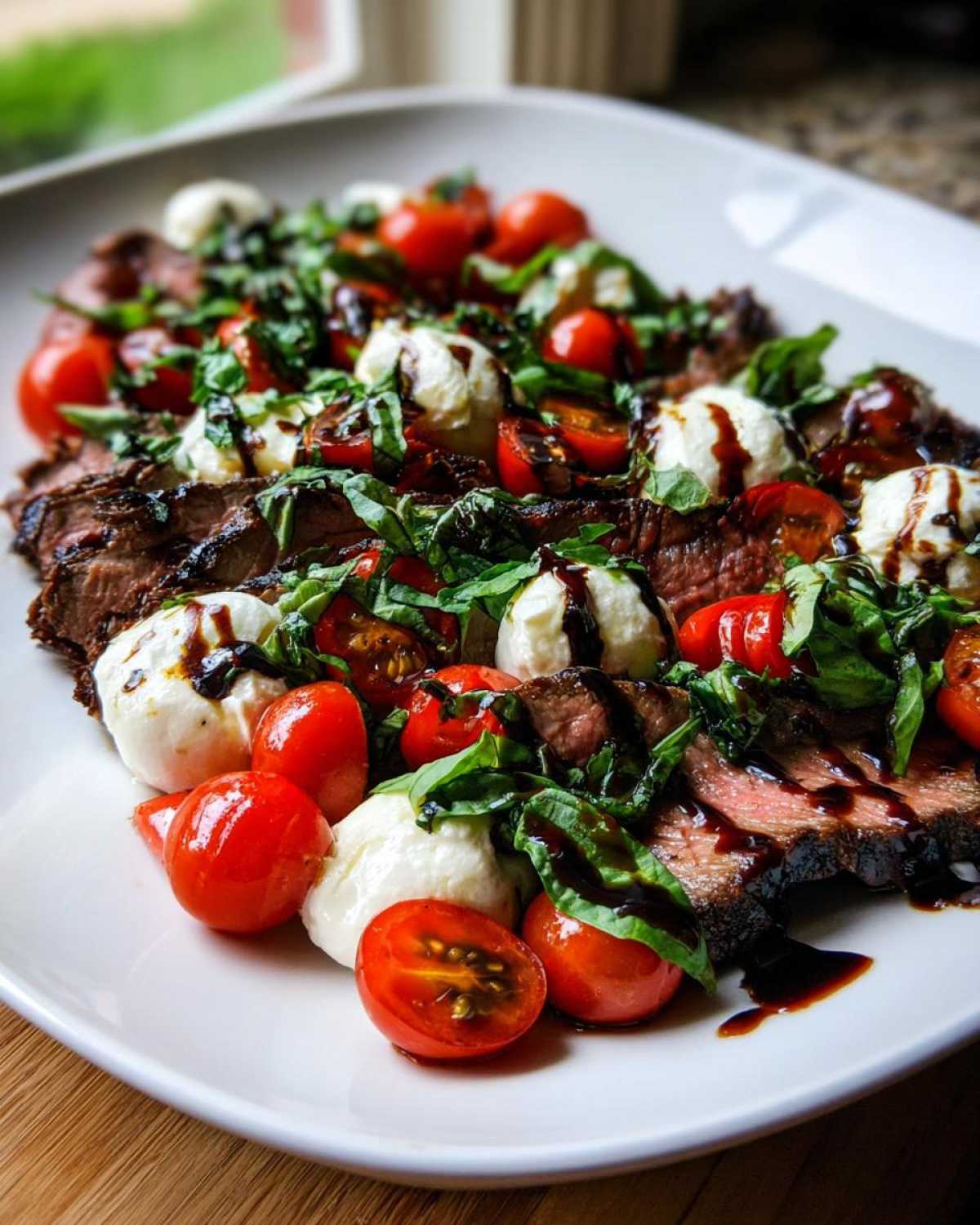Slices of Grilled Flank Steak Caprese topped with cherry tomatoes, mozzarella balls, basil, and balsamic glaze.