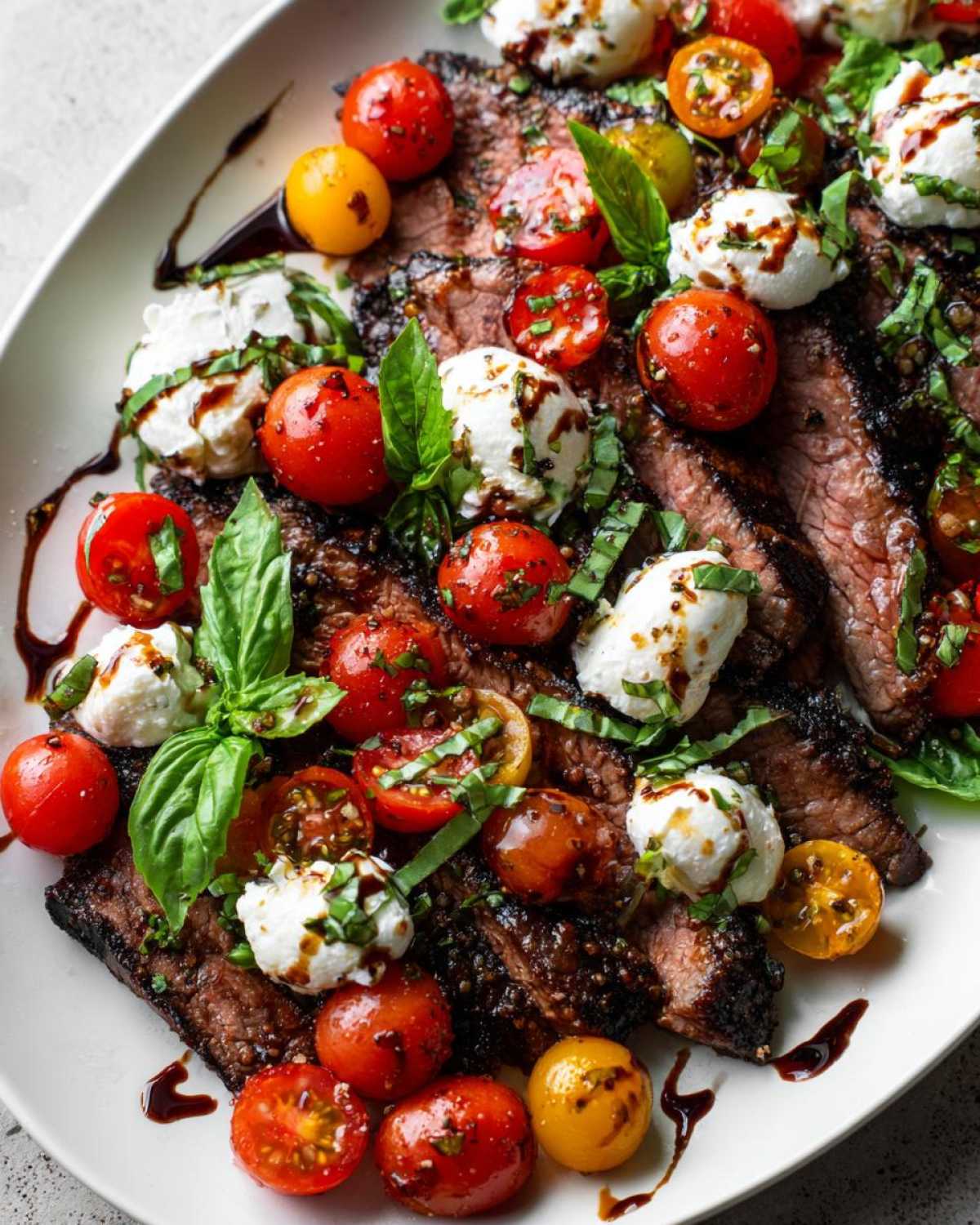 Close-up of sliced Grilled Flank Steak Caprese topped with cherry tomatoes, mozzarella balls, and fresh basil drizzled with balsamic glaze.