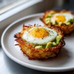 Close-up of a Hash Brown Avocado Egg Cup topped with a sunny-side-up egg and herbs.