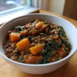 A close-up of a white bowl filled with Hearty Vegetable Lentil Soup featuring visible lentils, chunks of orange squash, and dark green kale.