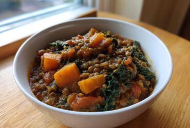 A close-up of a white bowl filled with Hearty Vegetable Lentil Soup featuring visible lentils, chunks of orange squash, and dark green kale.
