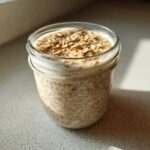 A glass jar filled with creamy Overnight Oats, topped with dry oats, sitting in bright sunlight.