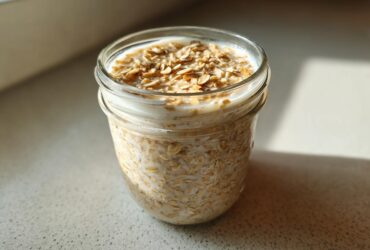 A glass jar filled with creamy Overnight Oats, topped with dry oats, sitting in bright sunlight.