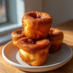 A stack of four golden brown, puffy Jordan Pond Popovers sitting on a small ceramic plate near a window.