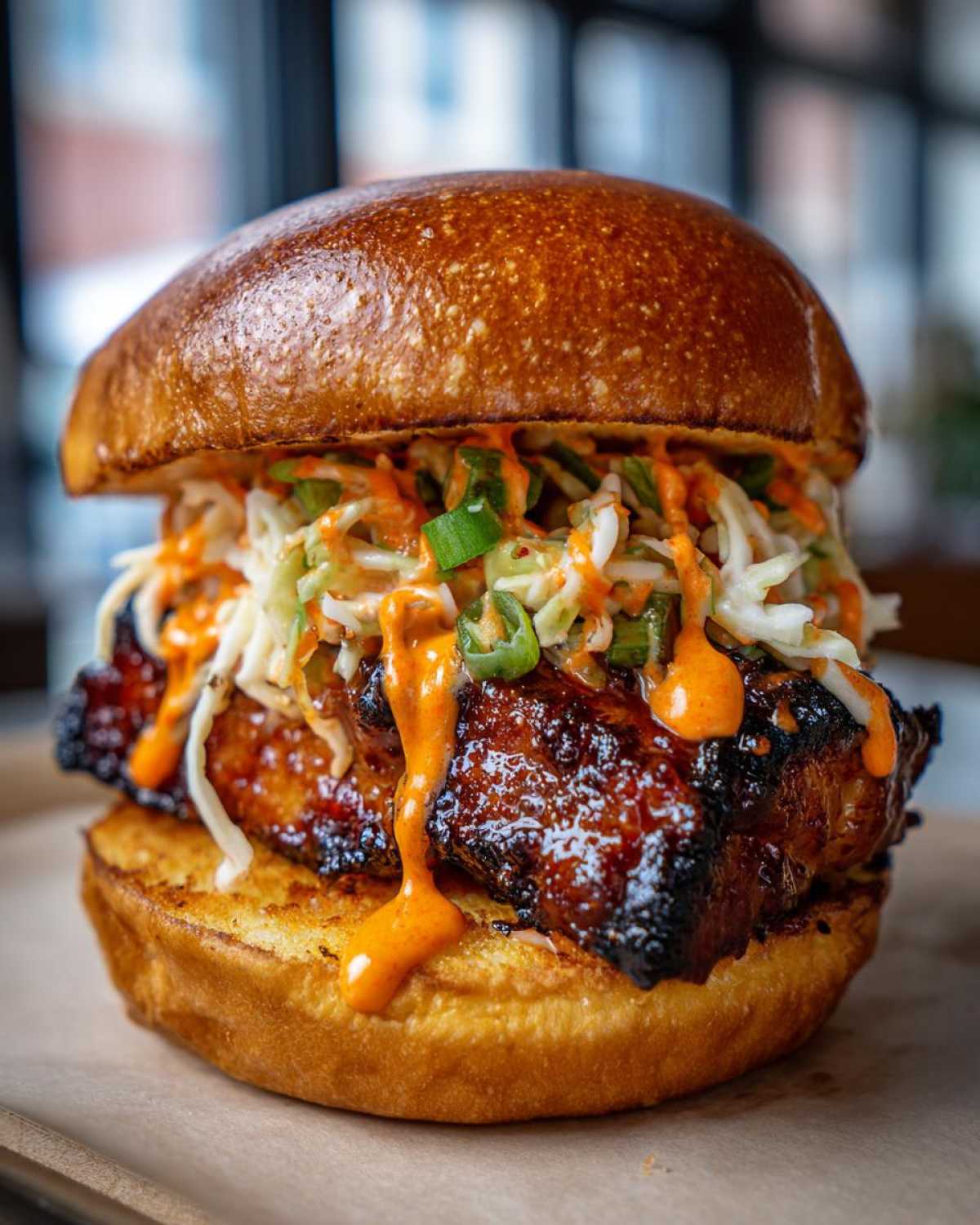 A close-up of a juicy KOREAN BBQ CHICKEN SANDWICH featuring glazed chicken, creamy slaw, and spicy orange sauce on a brioche bun.