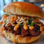 Close-up of a juicy KOREAN BBQ CHICKEN SANDWICH topped with creamy sauce and crunchy slaw on a toasted bun.