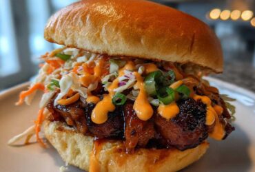 Close-up of a juicy KOREAN BBQ CHICKEN SANDWICH topped with creamy sauce and crunchy slaw on a toasted bun.