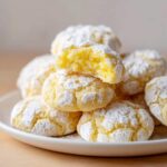 A stack of bright yellow Lemon Crinkle Cookies covered in powdered sugar, with one cookie broken open to show the soft interior.