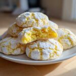 A stack of bright yellow Lemon Crinkle Cookies dusted heavily with powdered sugar, one cookie broken open to show the soft interior.