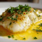 Close-up of a flaky white cod fillet swimming in bright lemon garlic butter sauce, topped with fresh parsley.