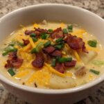 Close-up of a creamy bowl of Potato Soup topped generously with shredded cheddar cheese, crispy bacon bits, and sliced green onions.