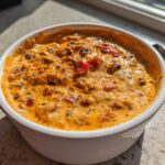 A close-up of a white bowl filled with rich, orange-hued Loaded Cowboy Queso, topped with spices and peppers.