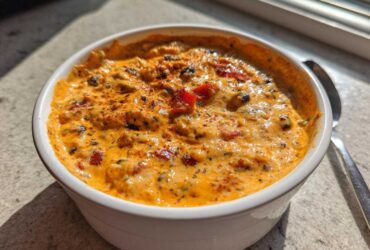 A close-up of a white bowl filled with rich, orange-hued Loaded Cowboy Queso, topped with spices and peppers.