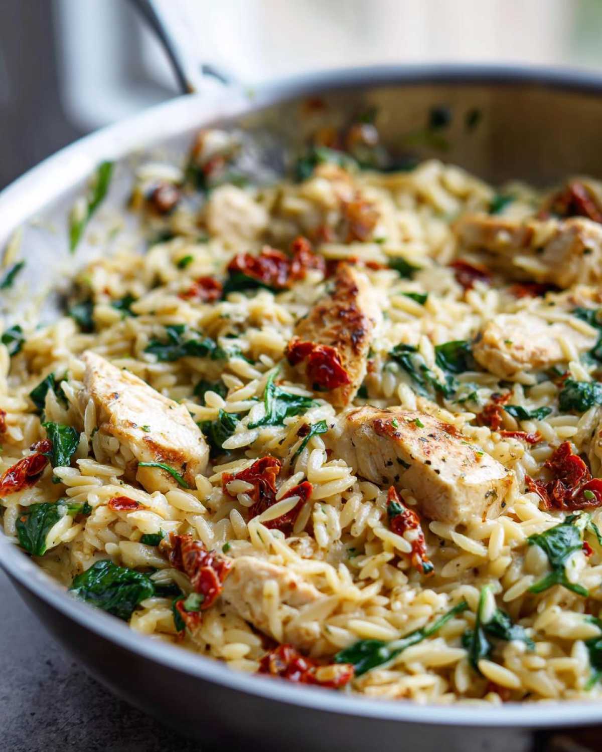 Close-up of creamy Marry Me Chicken Orzo mixed with spinach and sun-dried tomatoes in a stainless steel skillet.