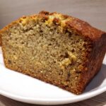 Close-up of a thick slice of moist Banana Bread showing a tender crumb and golden-brown crust, served on a white plate.
