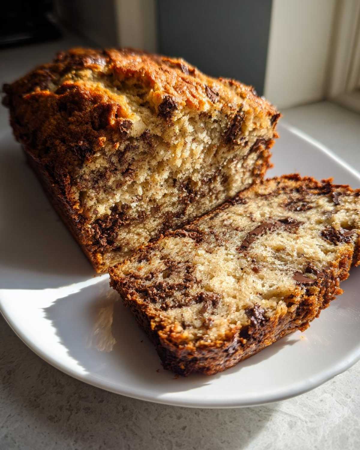 A moist Chocolate Chip Banana Bread loaf, partially sliced on a white plate, showing rich chocolate swirls.
