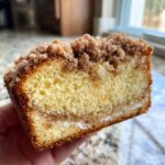 Close-up of a hand holding a slice of moist coffee cake showing a yellow crumb, cinnamon swirl, and thick streusel topping.