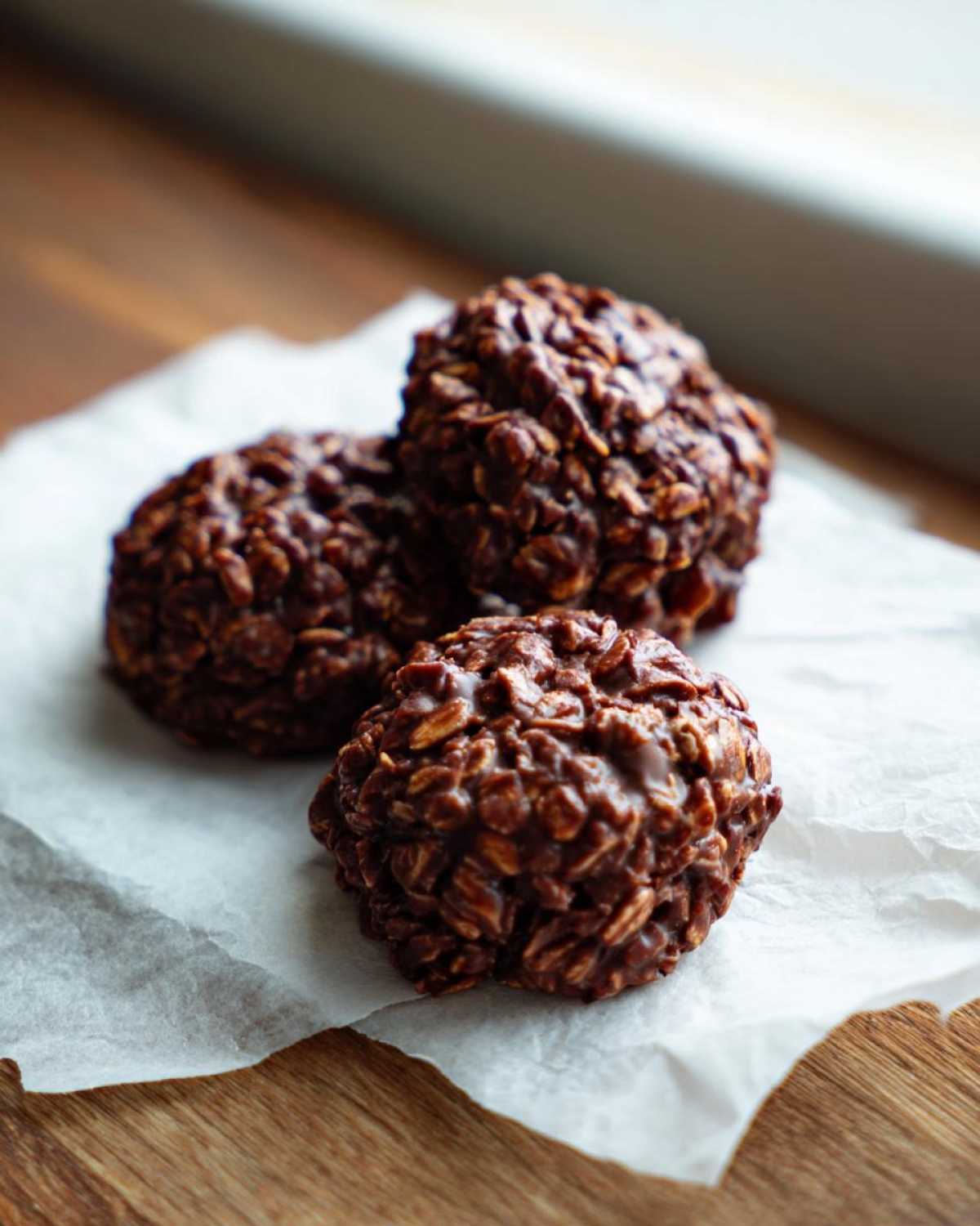 Three rich, dark No-Bake Chocolate Oat Cookies clustered on white parchment paper on a wooden surface.