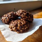 A close-up of three rich, dark No-Bake Chocolate Oat Cookies piled on a piece of parchment paper.