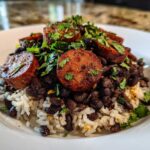 A close-up of a bowl featuring white rice topped with black beans, thick slices of seared sausage, and fresh cilantro.