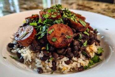 A close-up of a bowl featuring white rice topped with black beans, thick slices of seared sausage, and fresh cilantro.