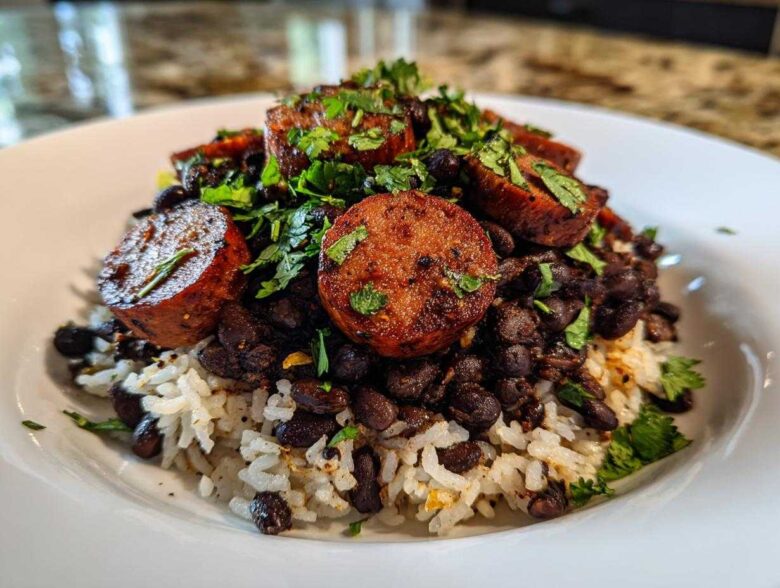 A close-up of a bowl featuring white rice topped with black beans, thick slices of seared sausage, and fresh cilantro.