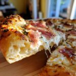 A close-up shot showing a slice being pulled from a Prosciutto Honey Burrata Pizza, highlighting the melted cheese stretch and toppings.