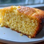 Close-up of a moist, golden slice of cornbread with a slightly browned crust, resting on a white plate.