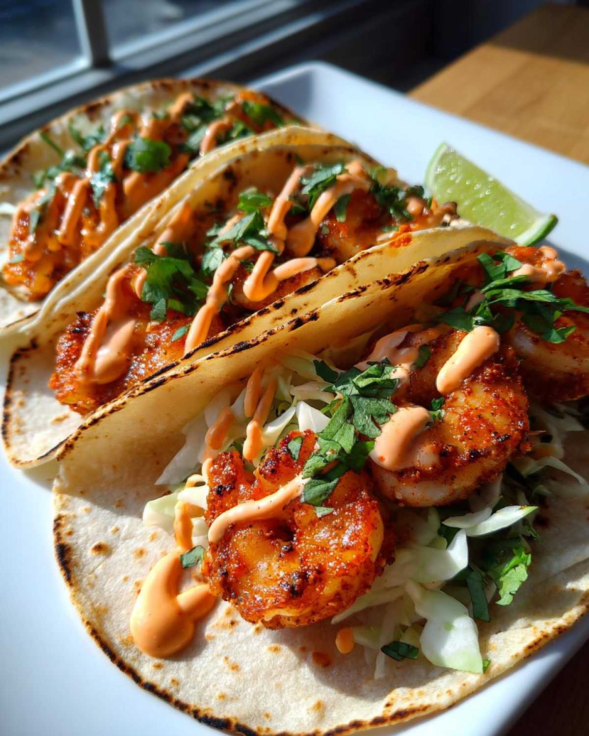Close-up of three Spicy Sriracha Mayo Shrimp Tacos topped with slaw, cilantro, and sauce, served with a lime wedge.