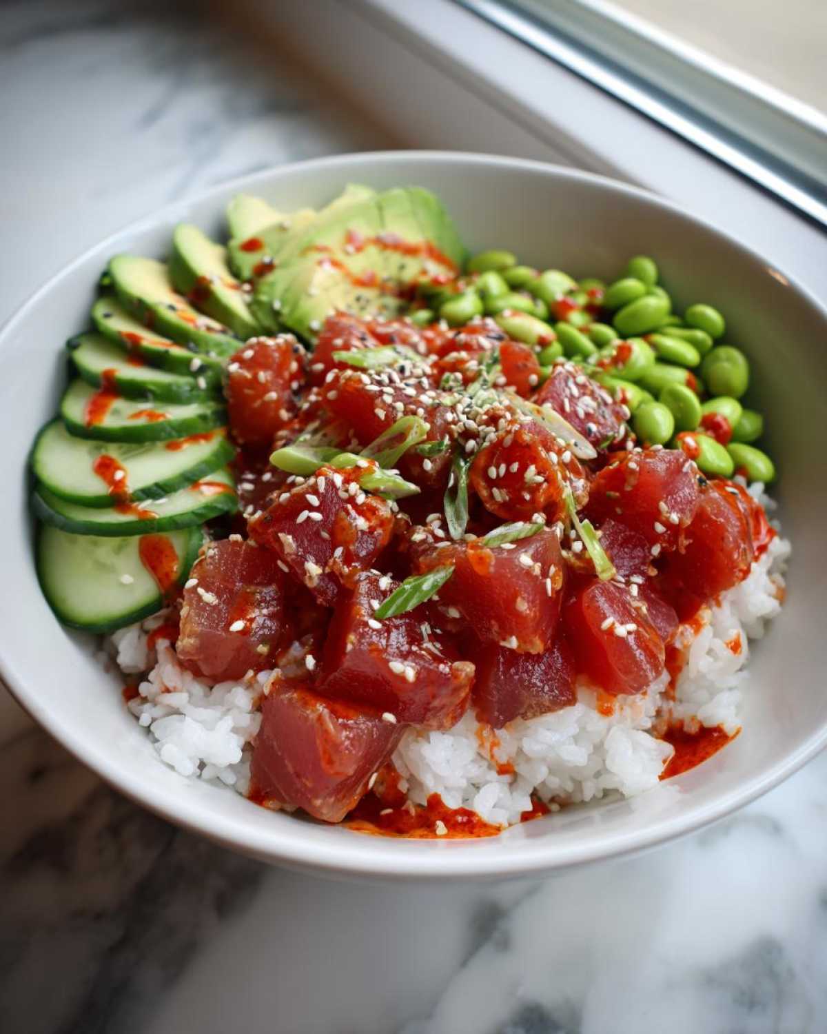 A vibrant bowl featuring marinated Spicy Tuna Bowl cubes over white rice, topped with sesame seeds, sliced avocado, cucumbers, and edamame.