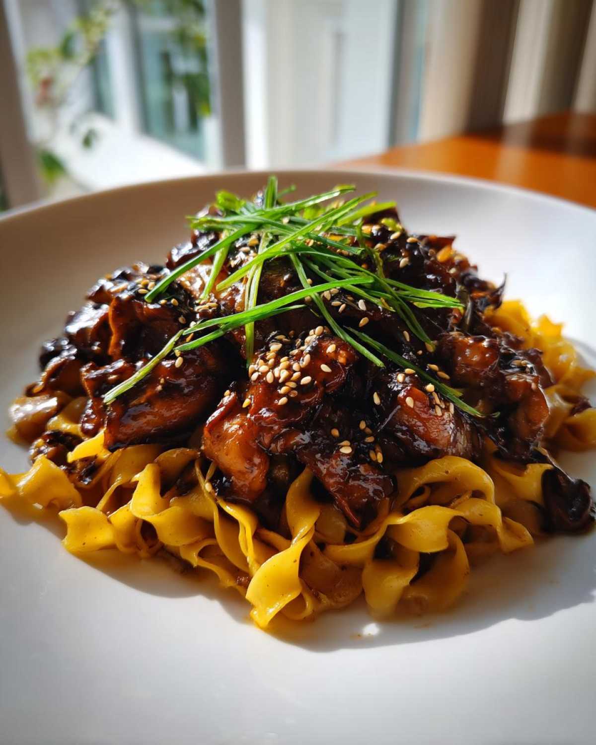 A serving of Sticky Garlic Chicken Noodles featuring dark, glossy chicken pieces over yellow egg noodles, garnished with sesame seeds and green onions.
