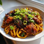 A close-up, appetizing shot of Sticky Garlic Chicken Noodles topped with sesame seeds and fresh green onions.
