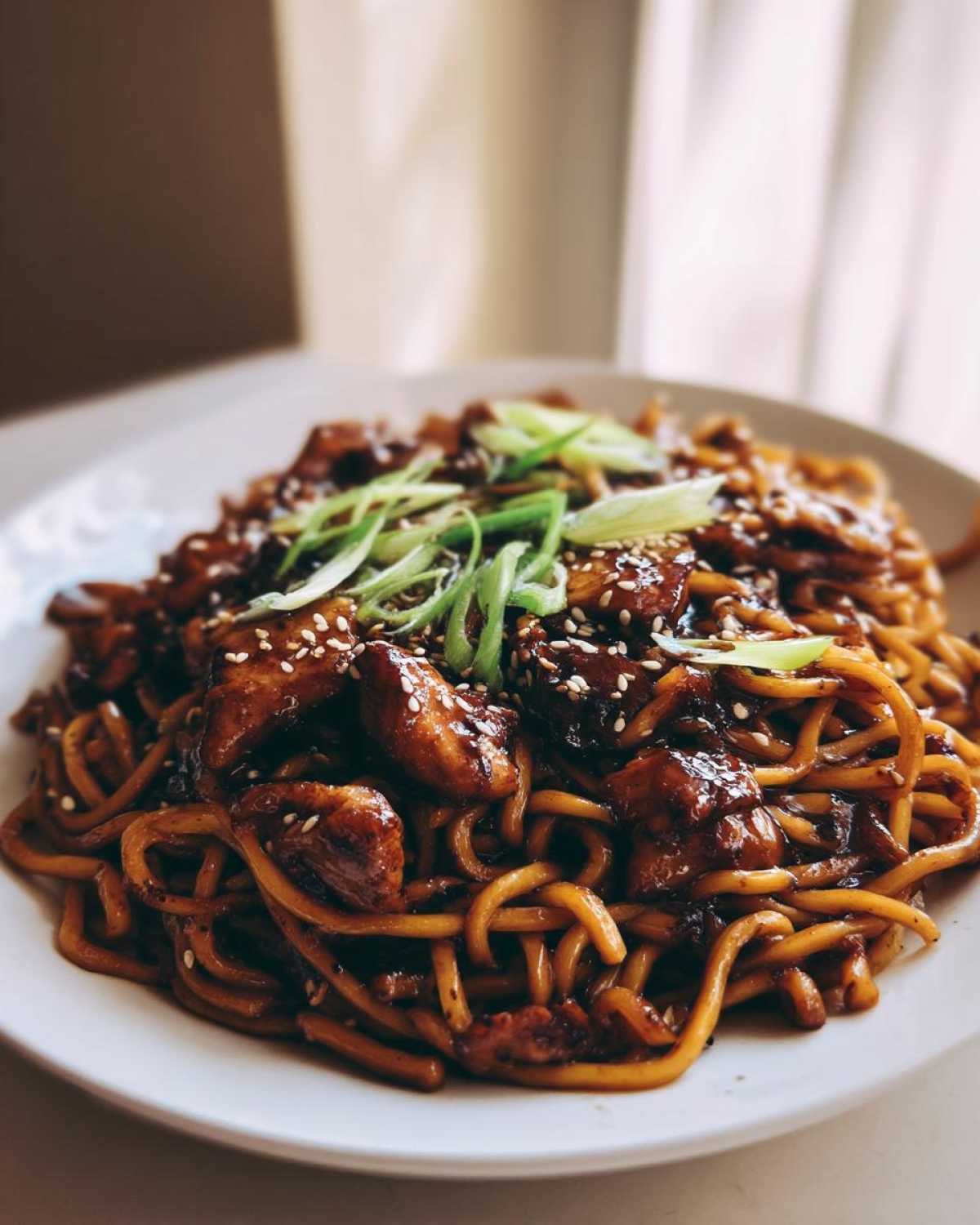 A close-up of Sticky Garlic Chicken Noodles coated in dark sauce, topped with sesame seeds and scallions.