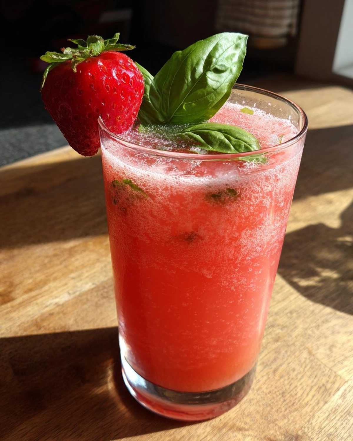 A tall glass of bright red Strawberry Basil Lemonade garnished with a fresh strawberry and basil leaves.