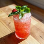 A tall glass of bright red Strawberry Basil Lemonade topped with crushed ice, fresh basil leaves, and a whole strawberry.