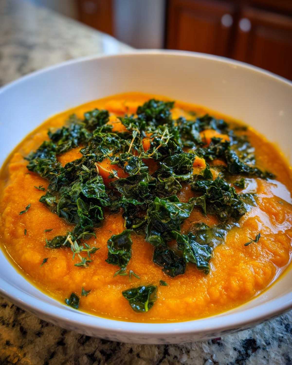 A close-up of a bowl of vibrant orange Sweet Potato Chowder Kale, topped with crispy green kale.